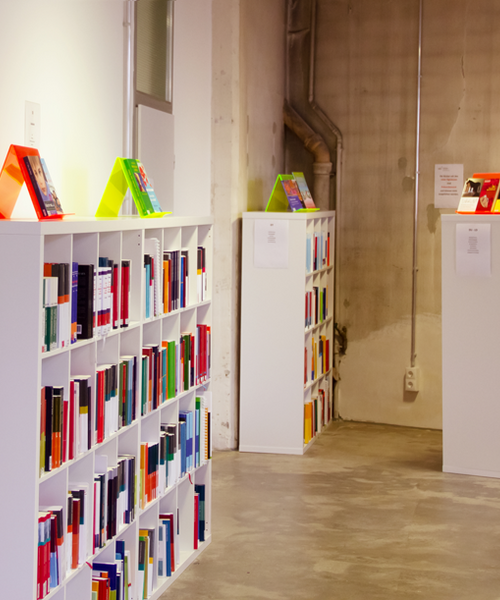 Regalreihen mit farbigen Büchern in einem modernen, hellen Raum mit Betonwand und klarer Beleuchtung.