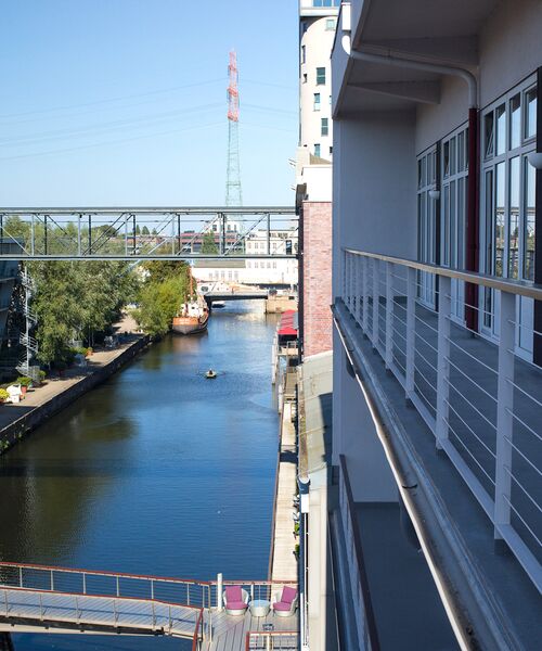 Blick von einem Gebäude auf einen Kanal mit klar blauem Wasser, flankiert von modernen Häusern und einer Brücke im Hintergrund. Ein Boot und ein Kajak sind auf dem Wasser zu sehen.