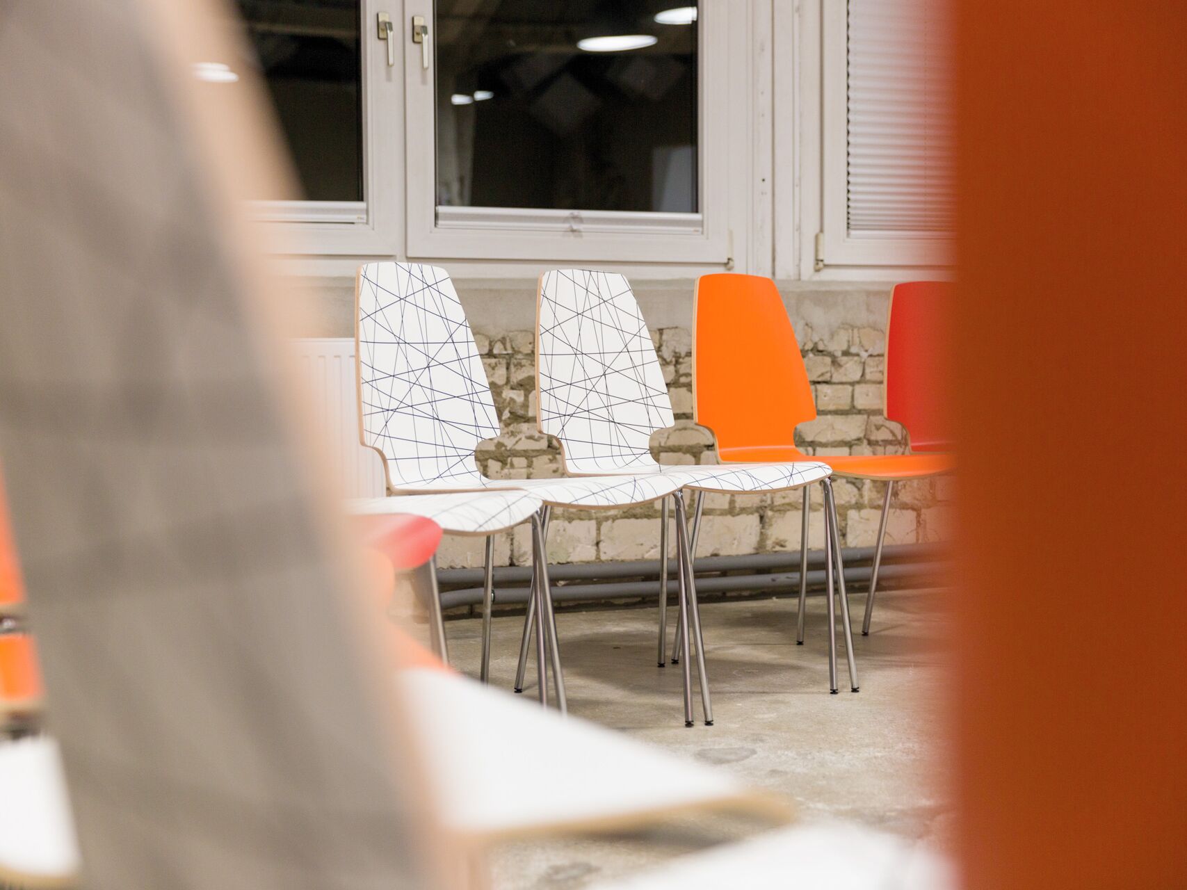 Mehrere moderne Stühle in Weiß und Orange stehen in einem Raum mit heller Fensterfront und Backsteinwand.