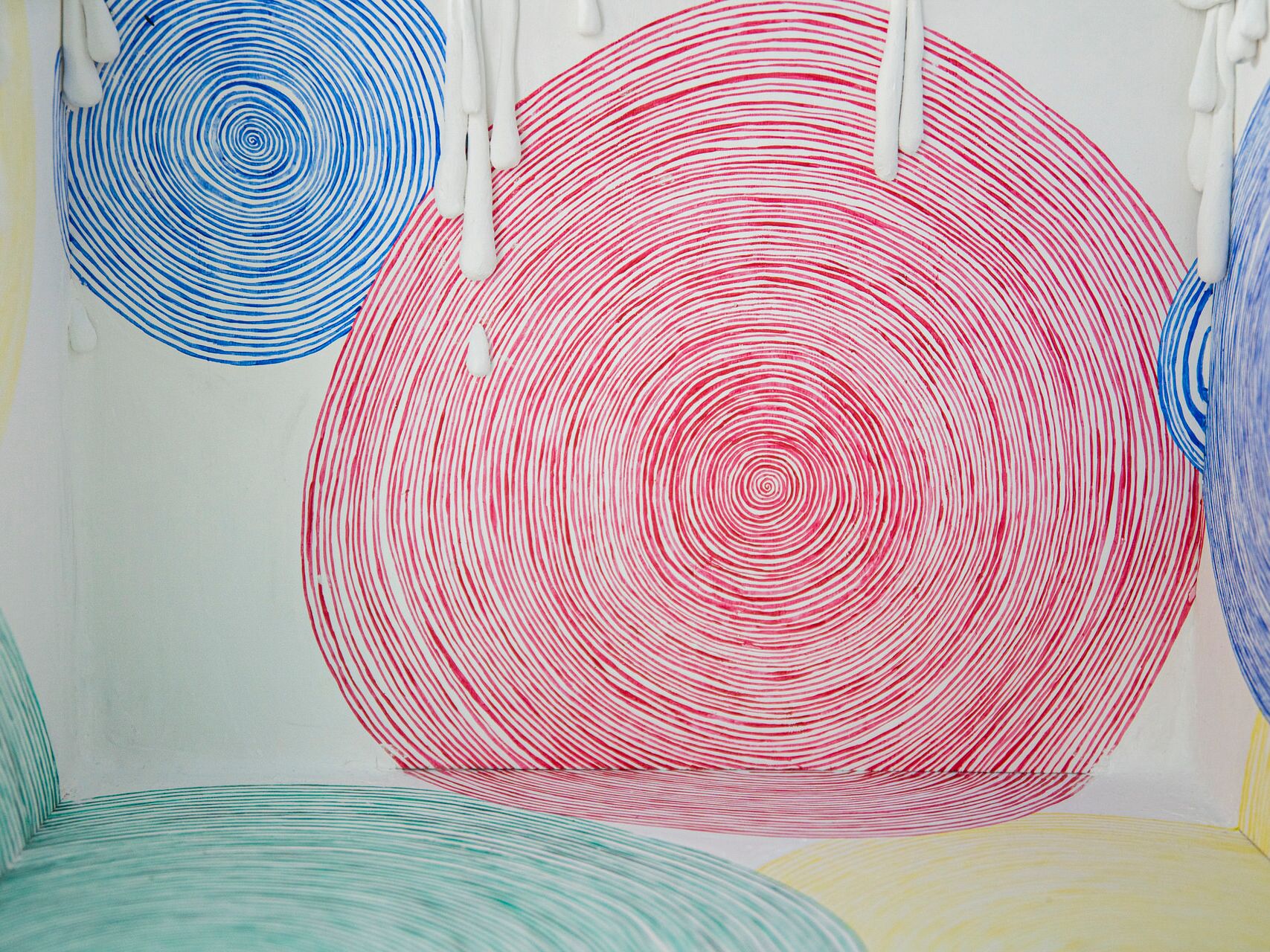 Bunte Kunstinstallation mit spiralförmigen Linien in Blau, Rot, Grün und Gelb auf einer weißen Fläche, von der weiße Wachstropfen herablaufen.