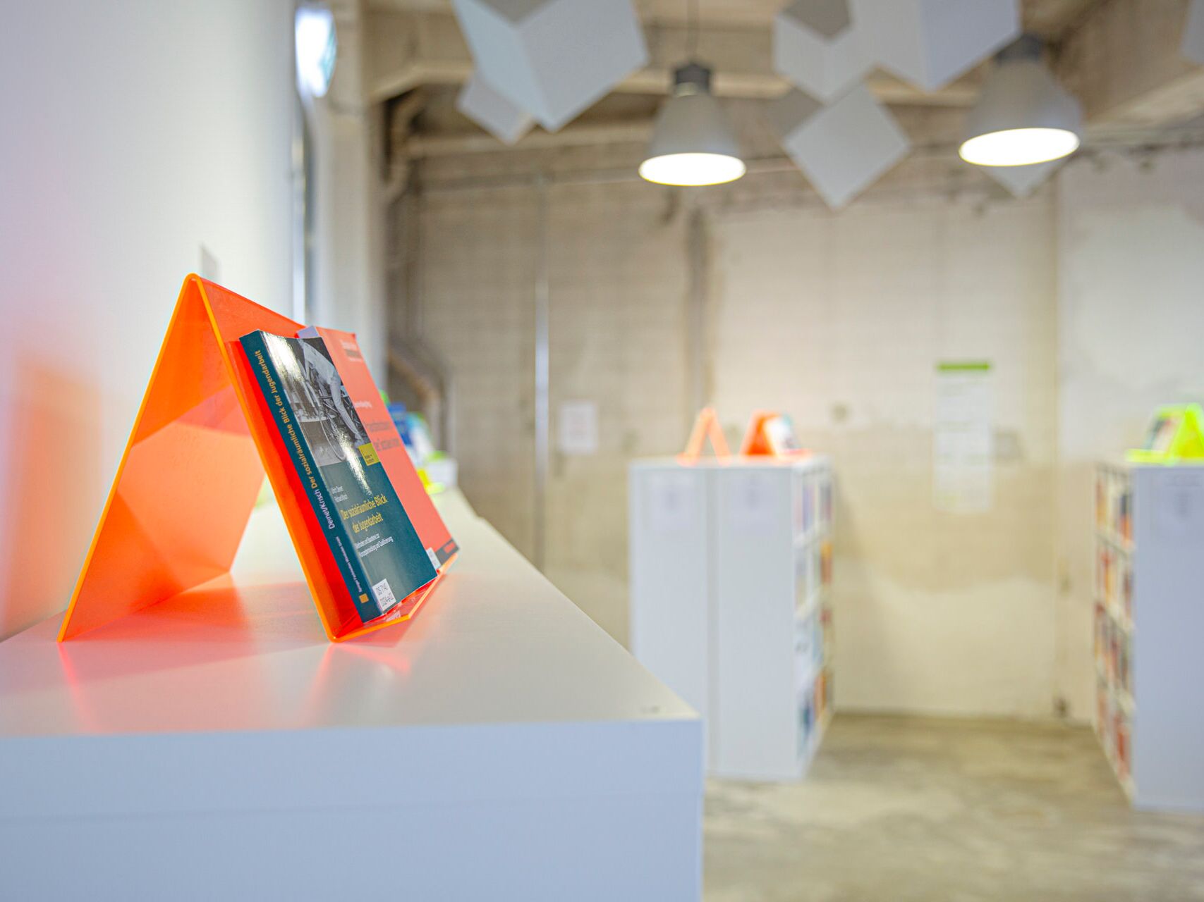 Heller Innenraum mit weißen Regalen und leuchtend orangefarbenen Buchständern, auf denen Fachliteratur präsentiert wird.