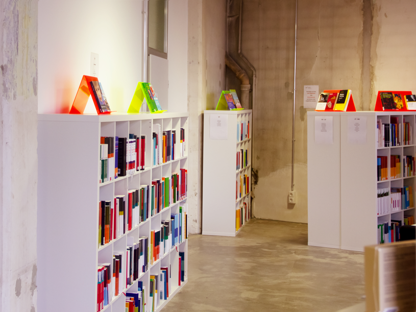 Regalreihen mit farbigen Büchern in einem modernen, hellen Raum mit Betonwand und klarer Beleuchtung.