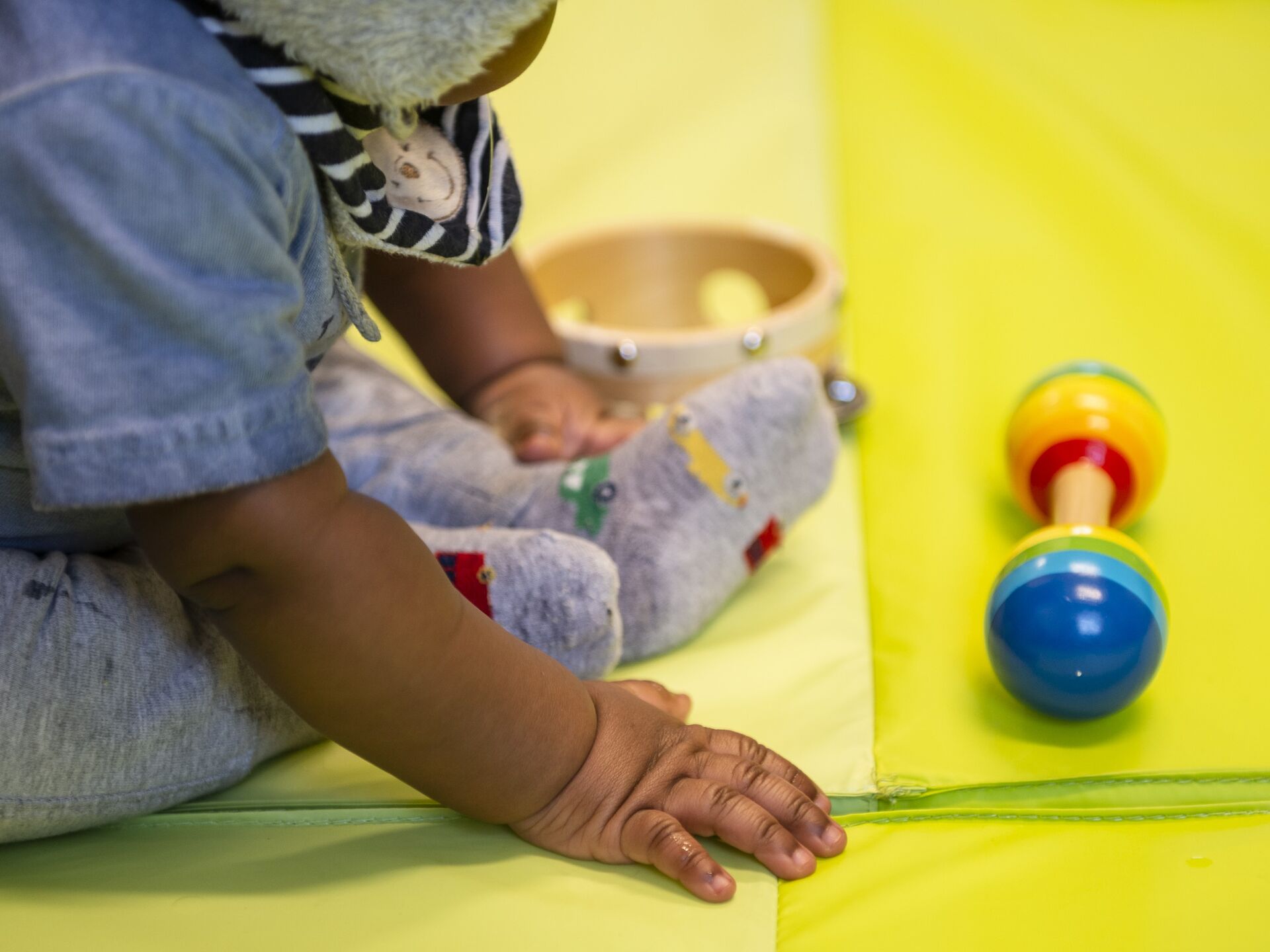 Kleinkind sitzt auf einer gelben Spielmatte, greift nach einem Spielzeug und hat eine kleine Trommel neben sich liegen.