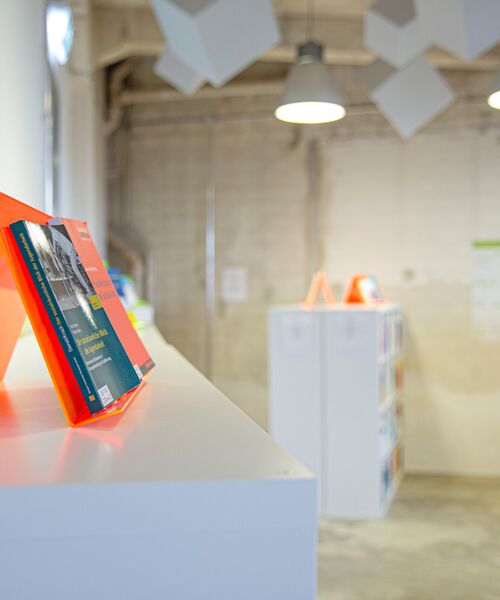 Heller Innenraum mit weißen Regalen und leuchtend orangefarbenen Buchständern, auf denen Fachliteratur präsentiert wird.