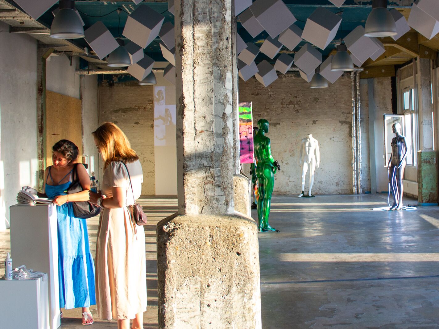 Menschen in einer hellen Kunstausstellung mit Betonpfeilern, farbigen Skulpturen und moderner Rauminstallation unter der Decke.